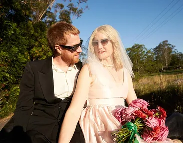 Bride in a sleeveless dress and veil holding flowers, sits next to man in a suit, both wearing sunglasses outdoors.