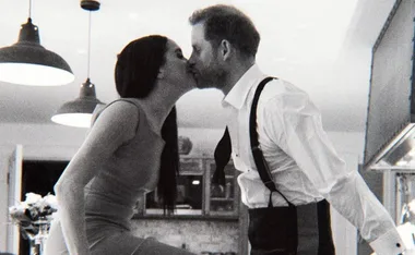 A couple kissing in a kitchen, with visible overhead lights and kitchen decor in the background.