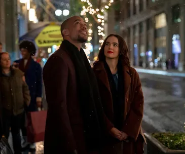 A couple stands on a snowy city street, surrounded by festive lights, gazing upward, with people and shops in the background.