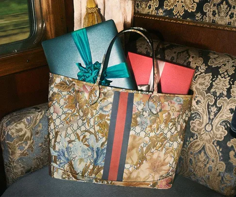 Gucci floral tote bag with gift-wrapped presents on a luxurious patterned seat, framed by a train window.