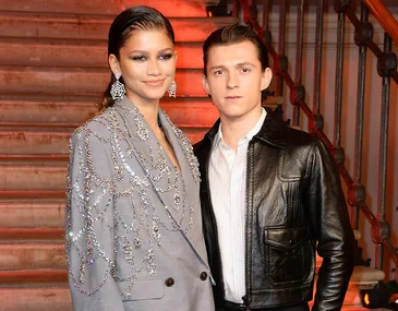 A stylish couple poses on a staircase; the woman wears a jeweled blazer, and the man a leather jacket.