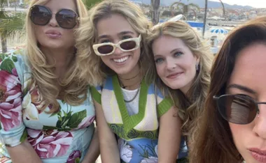 Four women smiling in sunglasses, posing closely outdoors with a scenic view in the background.