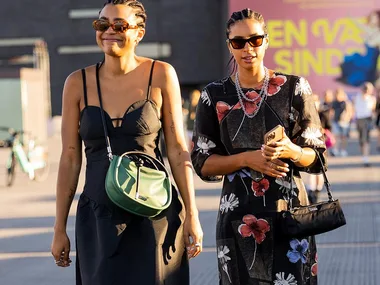 Two women wearing summer dresses and sunglasses, smiling on a street. One has a green bag, the other a floral dress.