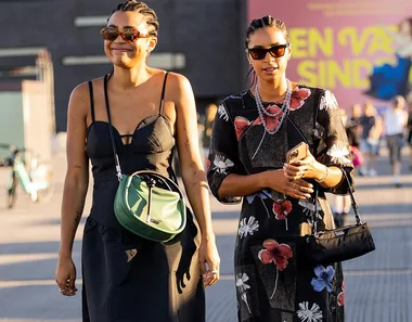 Two women wearing summer dresses and sunglasses, smiling on a street. One has a green bag, the other a floral dress.