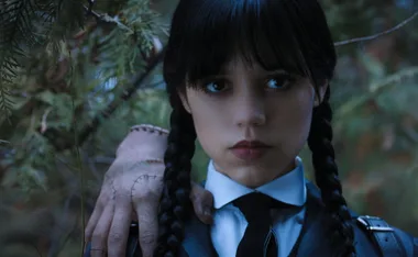 A young girl with braided hair and dark lipstick stands in a forest, with a stitched hand on her shoulder.