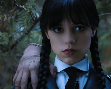 A young girl with braided hair and dark lipstick stands in a forest, with a stitched hand on her shoulder.