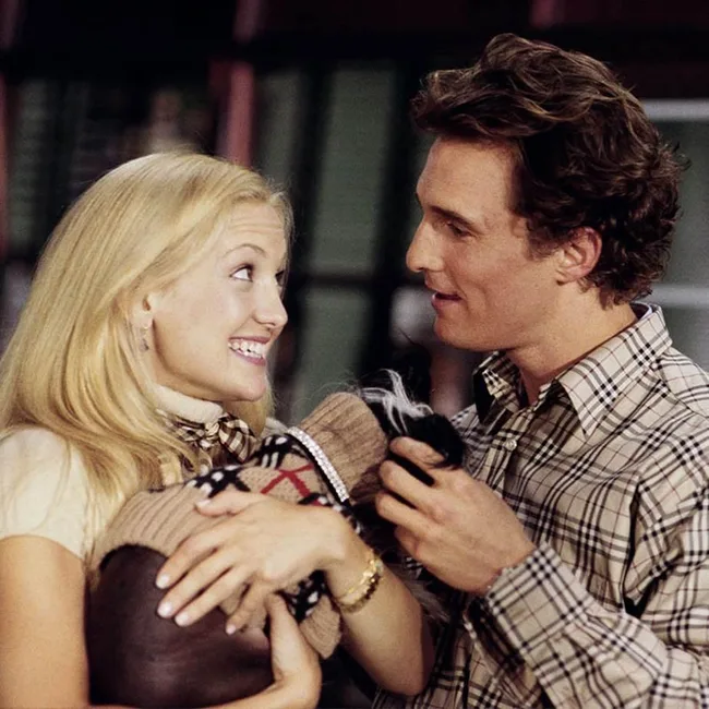 Couple in plaid embrace a dog wearing a sweater, smiling warmly at each other.