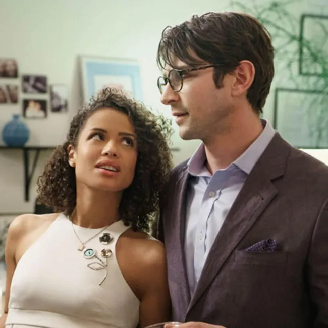 A woman in a white dress looks at a man in glasses and a suit in a room with photos on the wall.