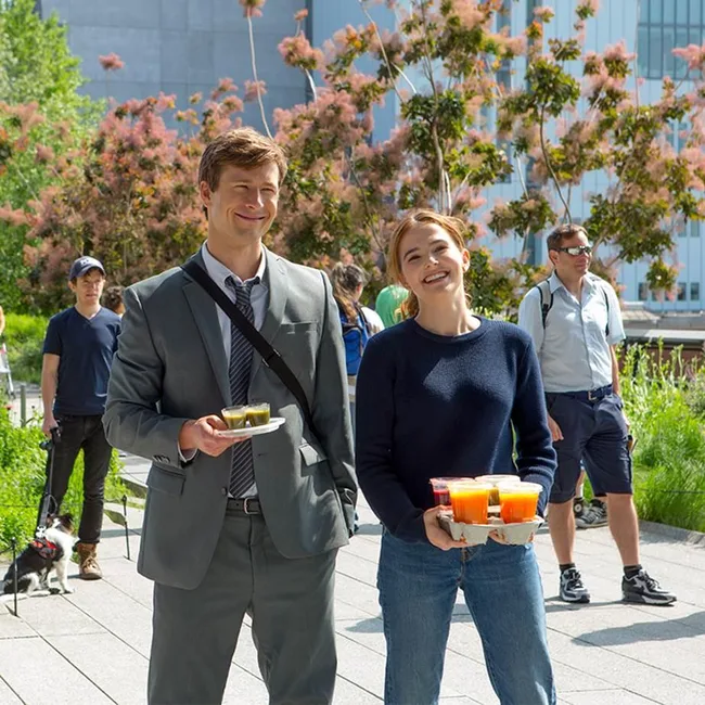 Young man in suit and woman in casual wear smiling while carrying drinks in a sunny park setting.