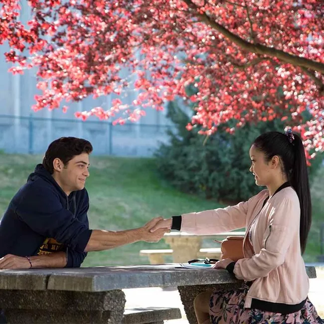 Two people shake hands across a picnic table under a tree with pink blossoms, in a park setting.