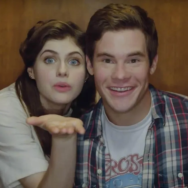 Two people smiling; one blowing a kiss. They appear happy, with casual clothing and wooden background.