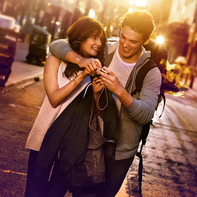 A young couple walking in a city street, smiling and looking at a smartphone as the sun sets behind them.