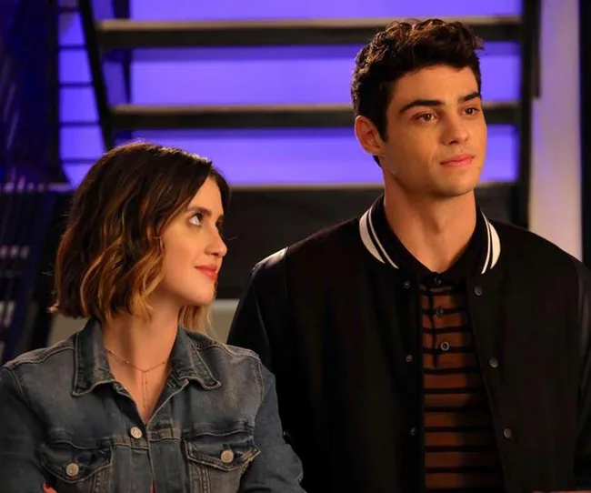 A woman in a denim jacket looks at a man in a black jacket with a striped shirt. Both stand in front of a purple-lit staircase.