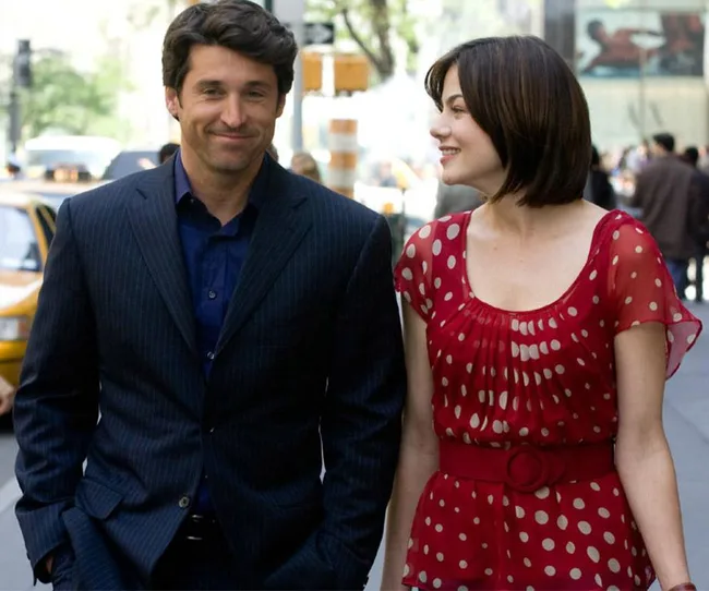 Man and woman walking on a city street, the man wears a suit, and the woman a red polka dot dress, smiling at each other.