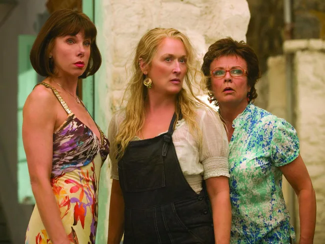 Three women stand close together, looking surprised, in colorful outfits in a sunny outdoor setting.