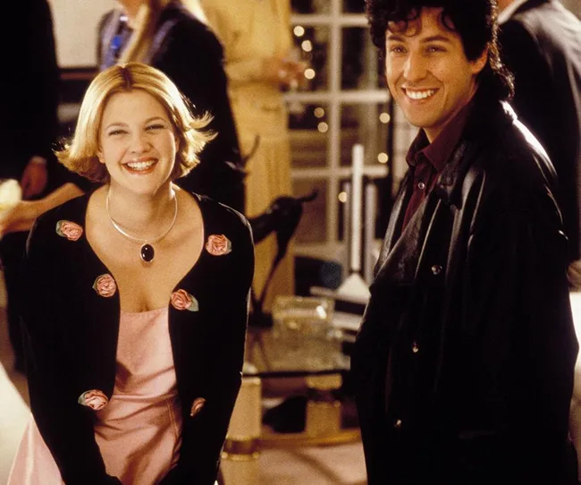 A woman in a floral cardigan and pink dress, and a man in a leather jacket, smiling at an indoor gathering.