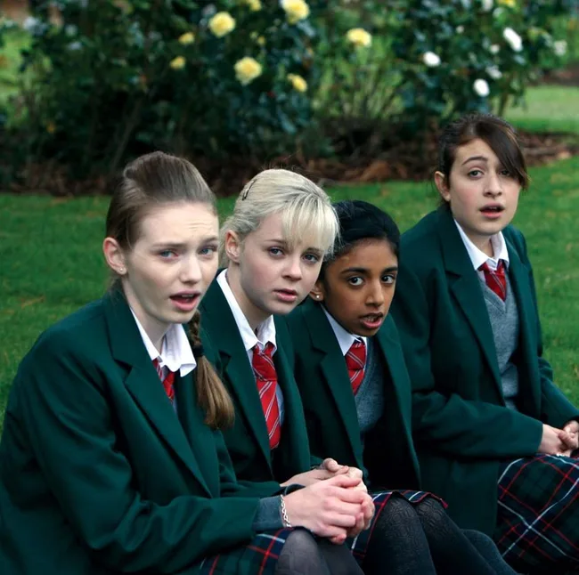 Four schoolgirls in green uniforms sit on grass, looking surprised. The background features yellow and white flowering bushes.