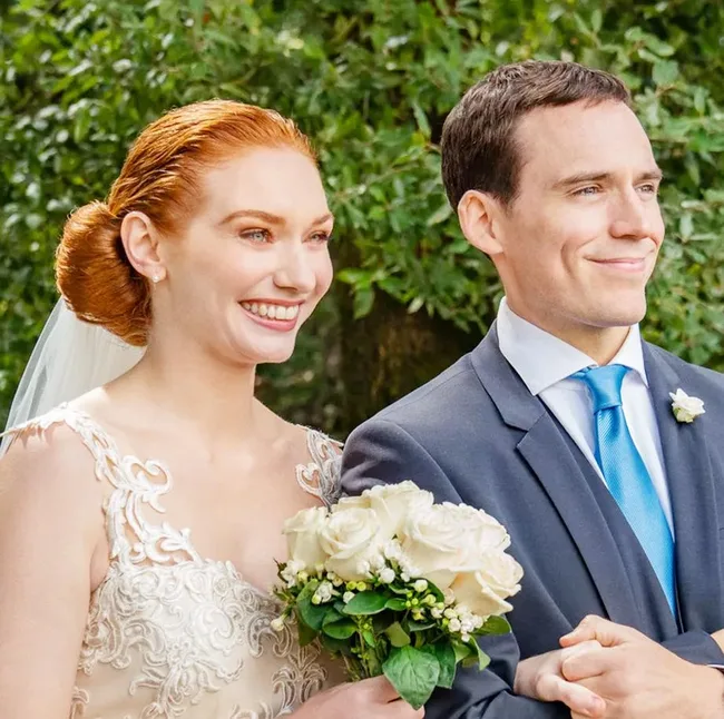 Wedding couple smiling, woman in lace dress holding bouquet, man in suit with blue tie, greenery background.