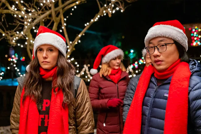 People wearing Santa hats and red scarves outside, with holiday lights in the background, looking serious.