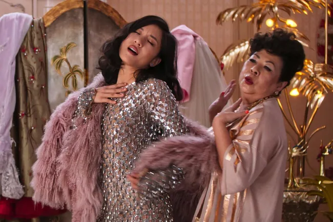 Two women posing dramatically in glamorous outfits in front of a decorative screen.