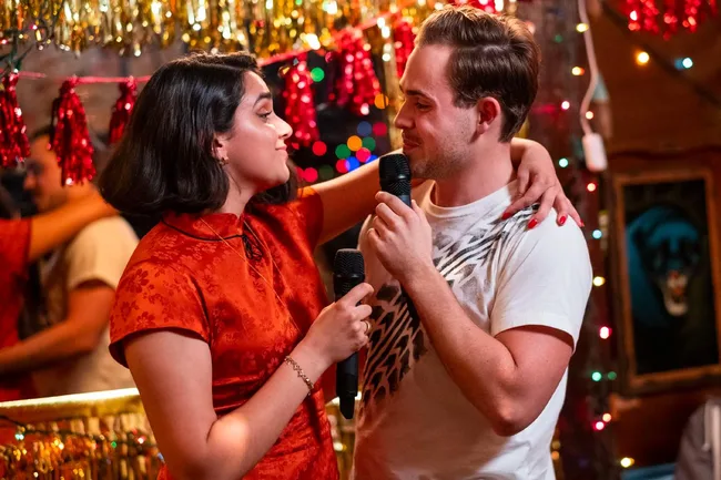 Two people holding microphones, smiling at each other, surrounded by festive lights and decorations.