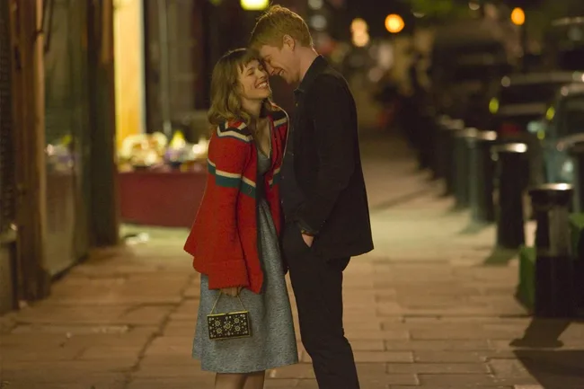 A couple leans in, sharing a joyful moment on a dimly lit street, with the woman wearing a red sweater and holding a purse.