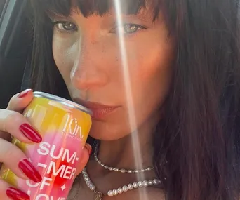 Woman with dark hair holds a can labeled "Kin Euphorics, Summer of Love," wearing a pearl necklace and red nails.