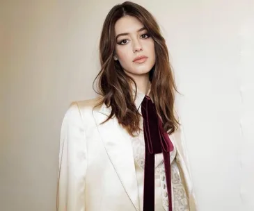 Young woman in a white suit with long brown hair and a maroon tie, posing against a plain background.