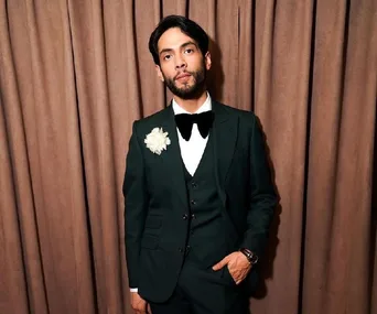 Man in a dark suit with a bow tie and floral lapel pin, standing in front of a curtain.