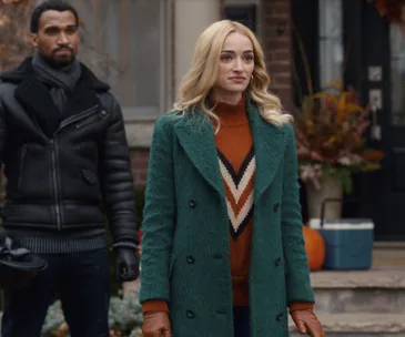 Woman wearing green coat with orange sweater stands outside a house, man in black jacket stands nearby.