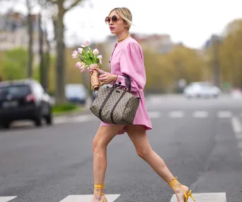 A woman in a pink dress and sunglasses walks with a bouquet of tulips and a designer handbag on a city street.