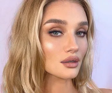 Blonde woman with glowy makeup, highlighting dewy skin and shimmering eye shadow, against a soft background.