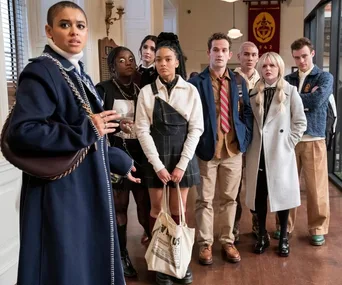 A group of young adults dressed in stylish, preppy outfits stands in a modern interior with serious expressions.