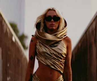 Singer in a gold dress and sunglasses walking on an outdoor path in a scene from a music video.