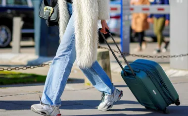 Person in jeans and fur coat walking with a teal suitcase on a city street.