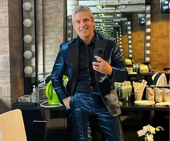 Man in shiny blue suit taking a mirror selfie in a dressing room with bulbs and various items on the counter.