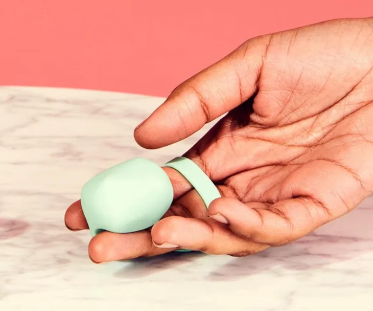 A hand holding a mint green finger vibrator with a loop, set against a pink and marble background.