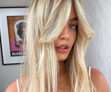 Blonde woman with long hair in front of an art piece, wearing a necklace and white top.