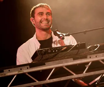 Performer smiling while playing keyboard at a concert with a microphone nearby.