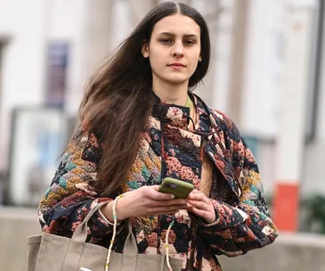 A person wearing a colorful jacket holds a phone and a Marc Jacobs tote bag while walking outside.