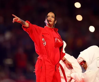 Rihanna performs at the Super Bowl halftime show in a red jumpsuit, pointing and surrounded by dancers in white.