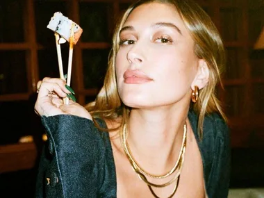 Woman with long hair holding sushi with chopsticks, wearing gold jewelry and a black outfit, looking at the camera.