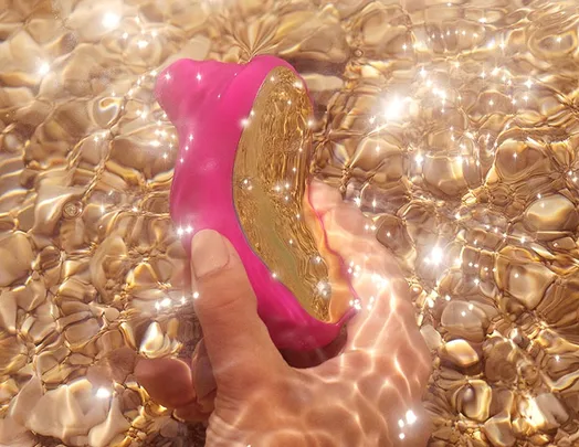 Hand holding a pink and gold vibrator underwater, with sunlight sparkling on the surface.