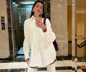 Person in a white oversized shirt and boots takes a selfie in a marble-floored elevator lobby.