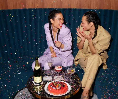 Two people with braided hair celebrate in a confetti-covered room, surrounded by drinks and desserts.