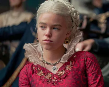 Character with braided blonde hair wearing a red dress, necklace with pendant, in a scene from "House of the Dragon."