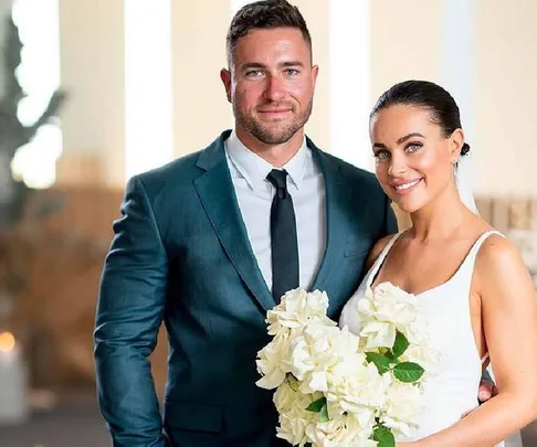 A couple from "Married at First Sight" stand smiling, the bride holding a bouquet of white roses.