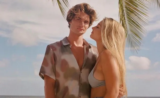 A couple stands on a beach under a palm tree; the woman looks at the man affectionately.