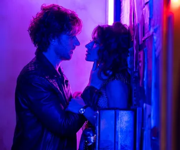 A couple in dramatic purple lighting, faces close, against a wall covered in posters.