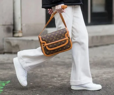 Person walking with a Louis Vuitton bag, wearing white pants and sneakers on a city street.